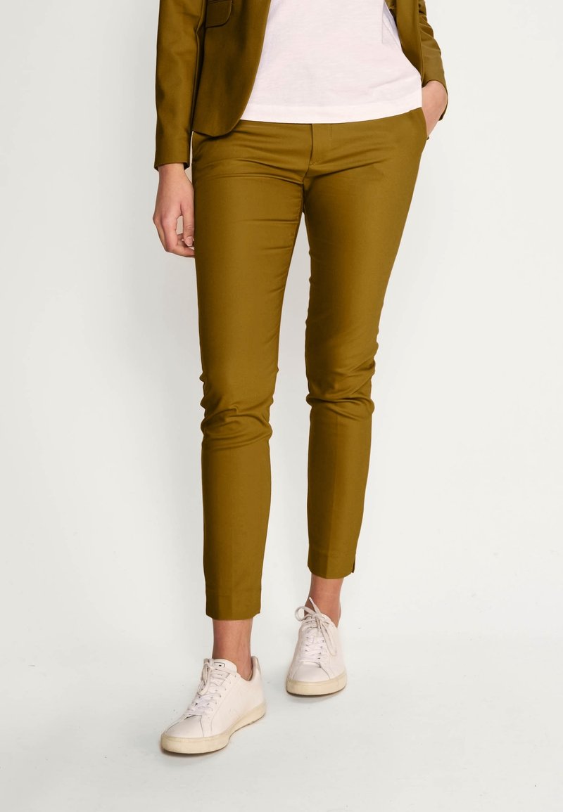 Personne portant un pantalon slim couleur moutarde, des baskets blanches et un blazer moutarde assorti avec une chemise blanche, debout avec une main dans la poche.