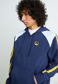 Navy hoodie with light blue shoulder accents, yellow side stripes, and a black and yellow hexagonal Nike logo. Features a front zipper and pockets.