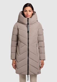 Beige polstret jakke med høj krave, chevron-syning, frontlynlås og logoaccent. Har sidelommer og et mellemlangt design.