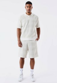 Polo texturé beige clair avec des manches courtes et un col doux, assorti à un short, porté avec des chaussettes blanches et des baskets.