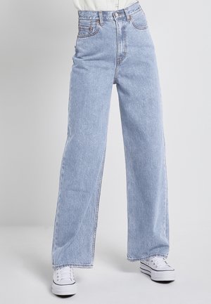 Jasnoniebieskie, szerokie dżinsy z wysokim stanem noszone z białymi trampkami na platformie na neutralnym, szarym tle.