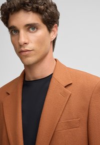Brauner strukturierter Blazer mit Reverskragen, einer einzelnen Brusttasche und einer klaren Silhouette, getragen über einem schwarzen Rundhals-Shirt.