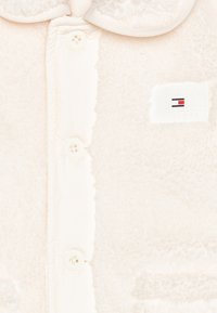 Primo piano di una giacca in pile color crema con bottoni, colletto e piccola patch rettangolare del logo Tommy Hilfiger sul petto.