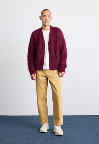 Burgundy fluffiga cardigan med framfickor, bärs över en vit skjorta, ihop med beiga byxor och ljusa sneakers på en blå matta.