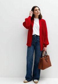 Röd stickad cardigan över en vit skjorta, kombinerad med blårandiga vida jeans och beige sneakers. Brunt handtag med strukturerad design.