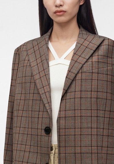 Personne portant un blazer à carreaux marron et gris par-dessus un haut blanc côtelé avec des bretelles croisées, visible des épaules à la taille sur un fond uni.
