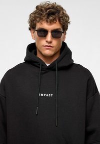Zwarte hoodie met een ruime pasvorm en een trekkoordcapuchon, voorzien van witte "IMPACT" tekst op de borst. Vergezeld van donkere zonnebril.