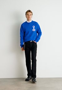 Blauwe sweatshirt met logo en witte accenten, zwarte slim-fit broek en zwarte schoenen; staande houding, een zwarte map vasthoudend.