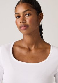 Λευκή ριμπ μπλούζα με φαρδιά λαιμόκοψη, απαλό ύφασμα και εφαρμοστή σχεδίαση. Το μοντέλο έχει απαλή επιδερμίδα και πλεξούδα στα μαλλιά.