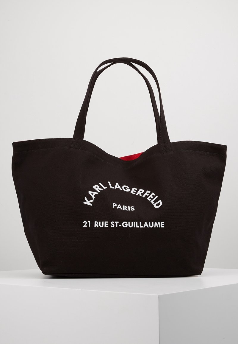 Sac cabas noir avec deux poignées, texte blanc "KARL LAGERFELD PARIS 21 RUE ST-GUILLAUME" sur le devant, doublure intérieure rouge légèrement visible.