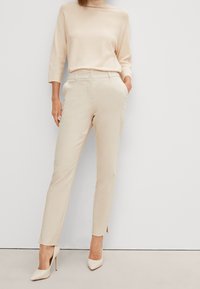 Top in maglia beige con collo alto e maniche a tre quarti, abbinato a pantaloni beige sartoriali con spacco laterale e bottone frontale.