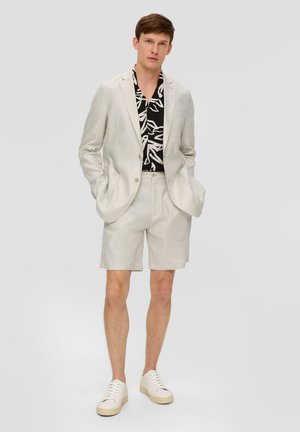 Helle Leinenanzug mit passender Shorts, getragen über einem schwarz-weiß gemusterten Hemd. Weiße Sneaker runden das Outfit ab.