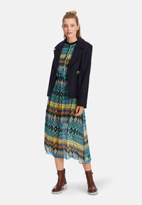 Langes, farbenfrohes Kleid mit Chevron-Muster in Blau, Grün, Orange und Gelb, kombiniert mit einem marineblauen Blazer und braunen Stiefeletten.