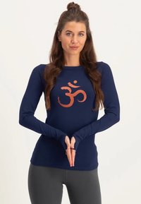 Mörkblå långärmad skjorta i mjukt tyg med en orange Om-symbol i mitten, och tumhål i ärmsluten.