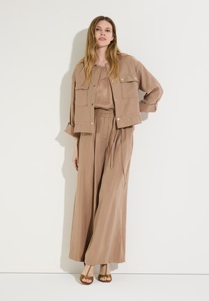 Frau steht vor weißer Wand, trägt eine lockere beige Jacke, passende weite Hosen und braune Schuhe mit offenem Zehenbereich, eine Hand in der Hüfte.