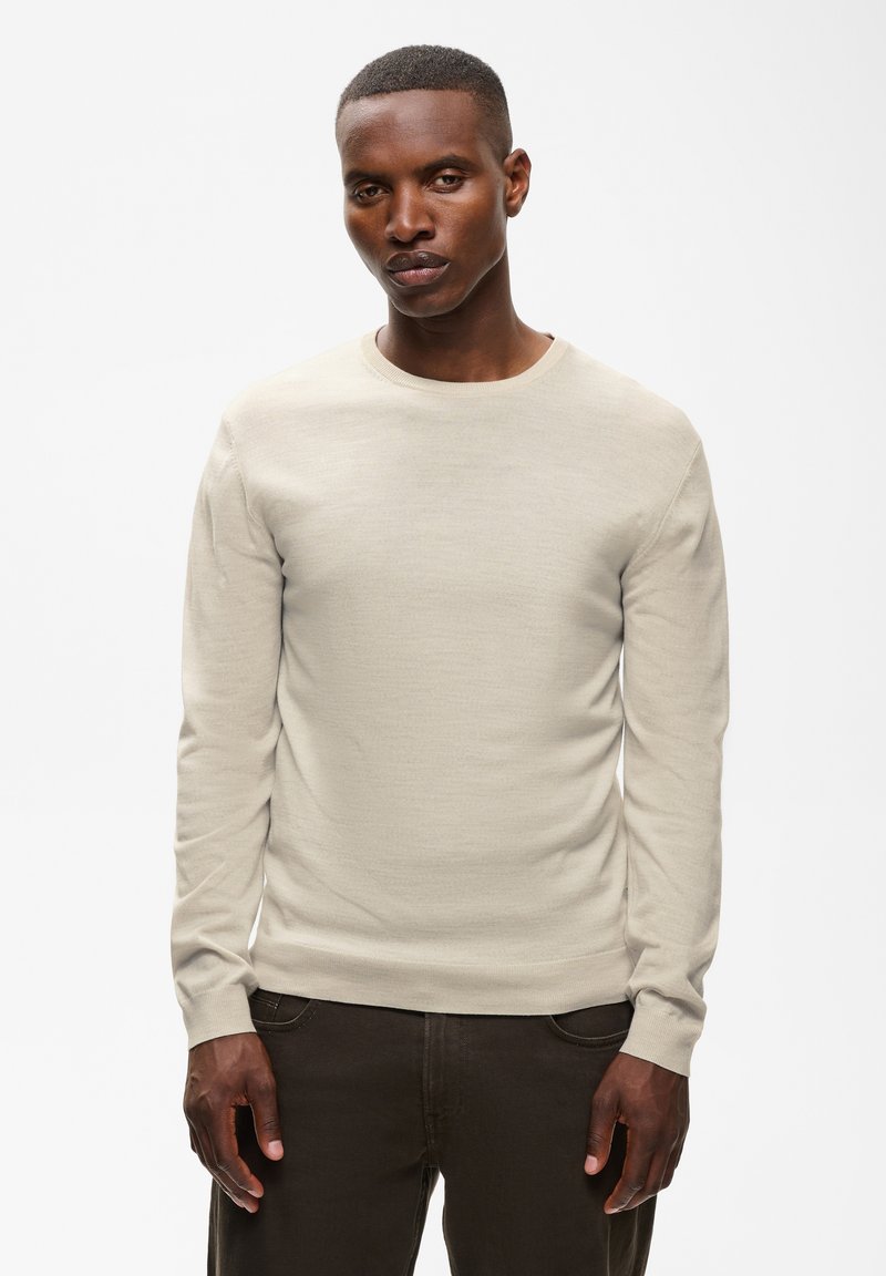 Homme portant une chemise beige clair à manches longues et un pantalon foncé, debout devant un fond blanc uni.
