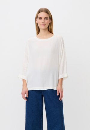 Frau mit langen blonden Haaren, die ein lockeres weißes Langarmshirt und weit geschnittene blaue Jeans trägt, steht vor einem schlichten weißen Hintergrund.