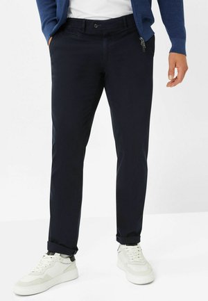 Mand iført mørkeblå straight-leg bukser, hvide sneakers, hvid skjorte og blå jakke, stående med hænder delvist i lommerne.