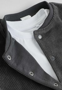 Tumehall puuvillane jakken, millel on nööbid ja valge T-särk all. Sile tekstuur ja nähtavad õmbluse detailid.