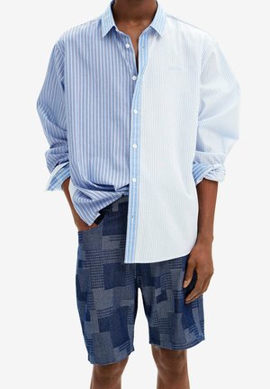 Homme portant une chemise à boutons rayée bleu clair et blanc à manches retroussées et un short bleu marine à motifs.