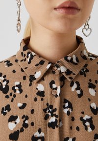 Καφέ πουκάμισο με μαύρες και λευκές λεοπάρ πιτσιλιές, υφασμάτινη υφή, κλασικό γιακά και κουμπί. Ορατά σκουλαρίκια σε σχήμα καρδιάς.