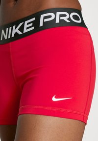 Shorts de sport rouges avec une large taille noire comportant "NIKE PRO" en blanc. Comprend un petit logo Nike blanc sur le bas gauche.