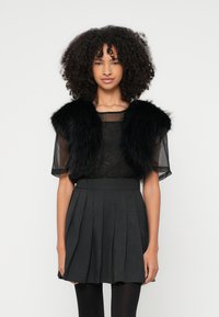 Bolero de pelo sintético preto sobre uma blusa preta transparente, combinada com uma saia plissada cinza escuro. Materiais texturizados com um design em camadas.