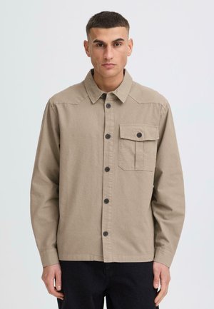 Mężczyzna ubrany w beżową koszulę na guziki z klapką na lewej piersi, stojący na tle jasnoszarym.