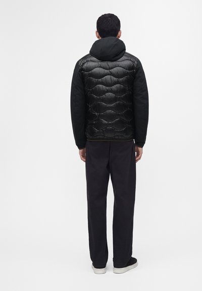 Homme debout, dos tourné, portant une veste matelassée noire avec capuche, un pantalon foncé et des chaussures noires aux semelles blanches sur un fond uni.