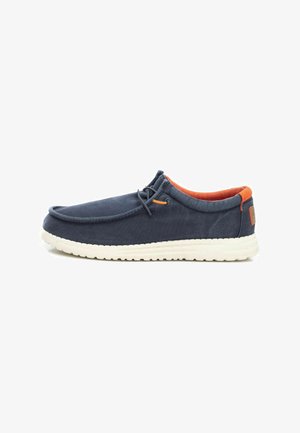 Zapato slip-on de lona azul marino con suela de goma blanca, interior naranja y un detalle decorativo de cordones en la parte superior.