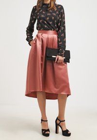 Svart blommig långärmad blus tillsammans med en högmidjad blush-pleated satin kjol. Modellen håller en svart kuvertväska och har på sig svarta plattformsskor.