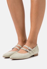 Ballerines en suède vert clair avec un bout pointu, deux brides à la cheville avec des boucles, et un petit talon en bois. Texture lisse avec un minimum de détails.