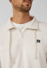 Pulóver color beige claro con cuello tipo zip, tejido texturizado, cordones y un pequeño parche con un logo en navy en el pecho.