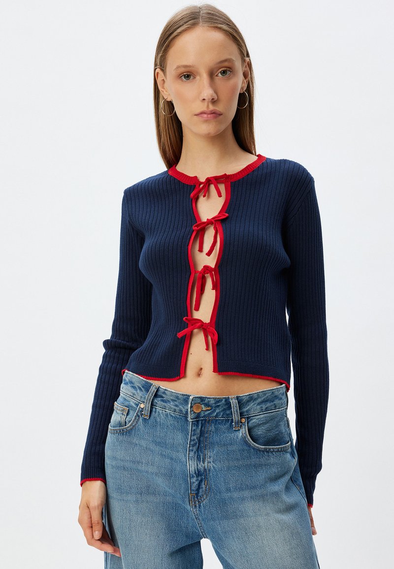 Cardigan a coste blu navy con accenti rossi, design corto, scollatura a chiave con lacci, maniche lunghe e vestibilità aderente. Jeans blu rovinati.