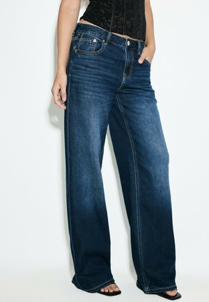 Kvinde iført mørkeblå jeans med vide ben og sort top, med den ene hånd i lommen og synlige fødder i sorte åbne sko.