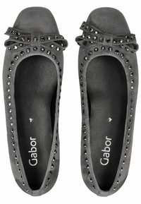 Mocasines de ante gris con punta redonda, adornados con tachuelas y un lazo en la parte superior. Interior negro con texto de logo. Textura suave en general.