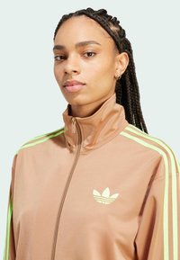 Braune Zip-up-Jacke mit grünen Streifen auf den Schultern, mit einem hellgrünen Logo auf der Brust. Glatte Textur und hoher Kragen.