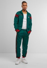 Trening teal cu accente roșii, jachetă cu fermoar complet și pantaloni cu talie elastică. Pantofii sport albi completează look-ul athletic.