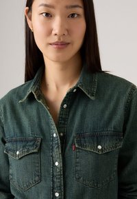 Groene denim overhemd met een knoopsluiting aan de voorkant, twee borstzakken en contrasterende stiksels. Zachte textuur met een merklabel op de linkerzak.