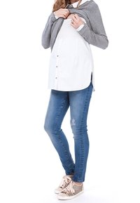 Outfit da gravidanza con una camicia bianca a bottoni, un cardigan grigio, jeans skinny blu e sneakers grigie con dettagli e lacci beige.