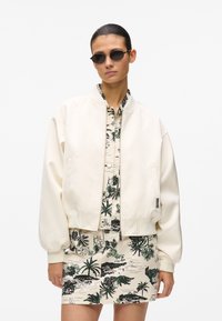 Blouson aviateur crème avec manches retroussées, porté sur une chemise à motifs arborant des palmiers verts et noirs sur fond beige.