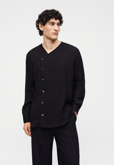 Νεαρός άνδρας με σγουρά μαλλιά που φοράει μαύρο ασύμμετρο πουκάμισο με κουμπιά μακρυμάνικο και μαύρο παντελόνι, στέκεται με το ένα χέρι στην τσέπη.