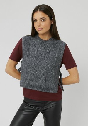 Gilet en laine grise tricoté avec un motif en relief, doté d'un ourlet côtelé et de liens sur les côtés, porté sur une chemise à manches courtes bordeaux.