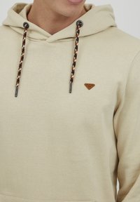 Felpa beige con tasca a marsupio anteriore, presenta cordini intrecciati neri e arancioni, e un piccolo logo marrone sul petto.