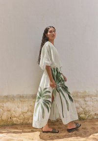 Long white dress with palm leaf print, cinched waist, and side slit. Model wears dark green slide sandals, standing against a textured wall.