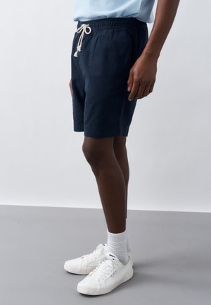 Άνδρας που φοράει σκούρο μπλε σορτς με κορδόνια, λευκές κάλτσες πάνω από τον αστράγαλο και λευκά αθλητικά παπούτσια, στέκεται μπροστά σε απλό γκρι και λευκό φόντο.