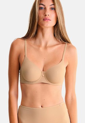 Femme aux cheveux longs portant un soutien-gorge beige sans couture avec armatures et une culotte taille haute assortie, sur fond blanc uni.
