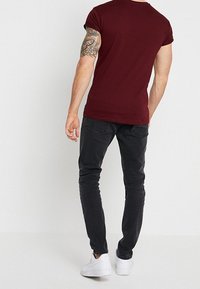 Camiseta ajustada color marrón con mangas enrolladas, combinada con pantalones slim-fit gris oscuro y zapatillas blancas. Tatuaje visible en el brazo.
