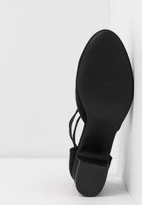 Chaussure à talon noire pour femme reposant verticalement sur un coin blanc, montrant une semelle lisse et le dessous texturé du talon.