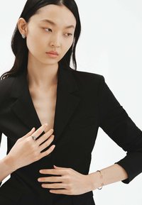 Blazer negro con un profundo escote en V, dos bolsillos delanteros y una tela texturizada. Manos adornadas con un anillo de plata y una delicada pulsera con acentos rosas.